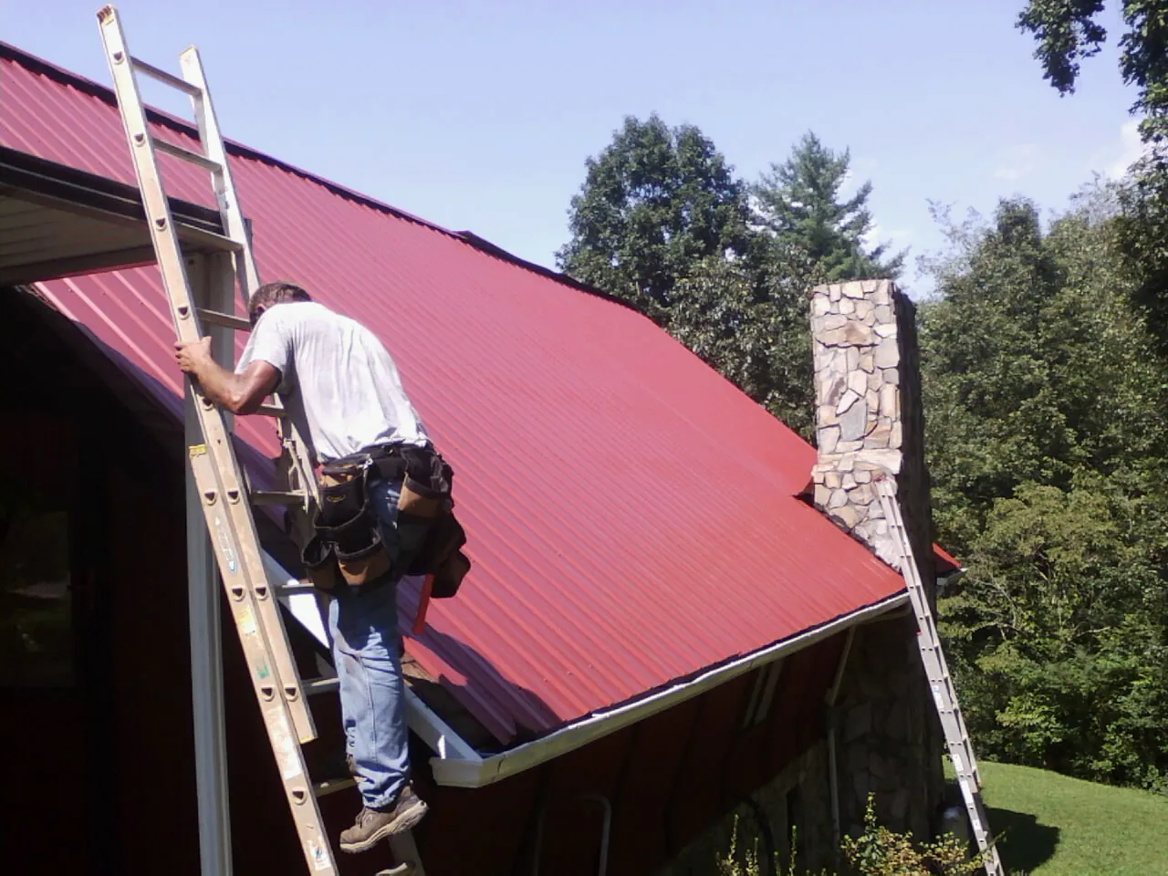 Roofer on a ladder beside a red metal roof and stone chimney — Soffit & Fascia Repair by Roofing Brothers in Brecksville
